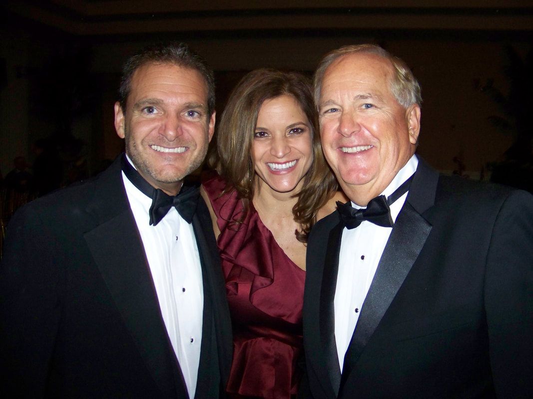 Three smiling people in formal wear pose for a photo. Woman in a red dress smiles between two men in tuxedos.