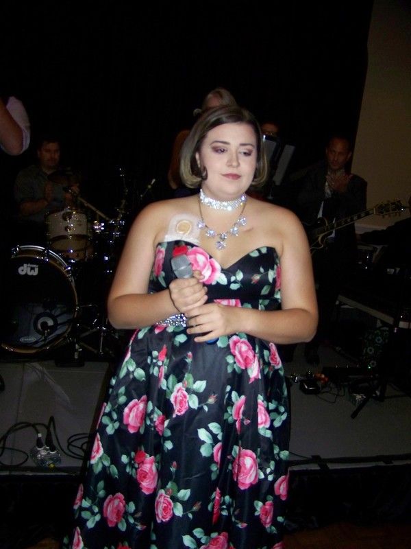 Woman in a black rose-print dress sings into a microphone with a band behind her at a dark event.