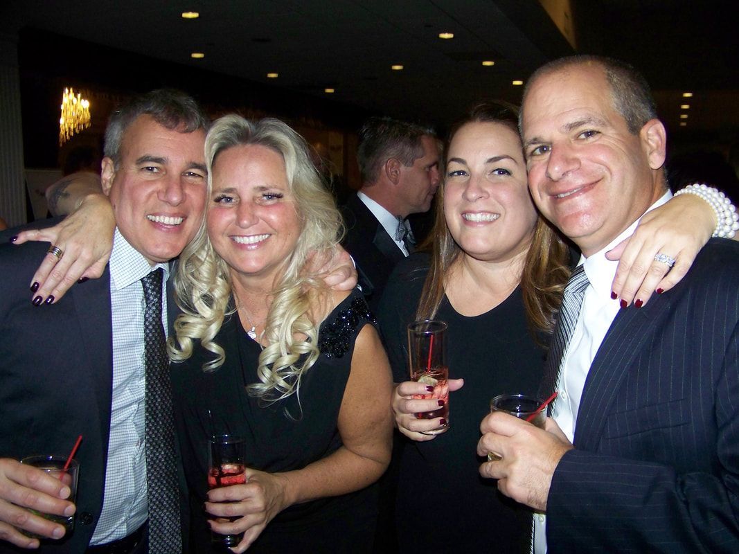 Four people smiling, holding drinks, arms around each other. Inside, dressed in formal attire.