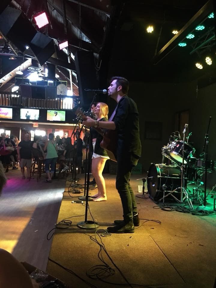 Band performing on a stage. Man singing with guitar, woman next to him. Venue interior, audience in background.