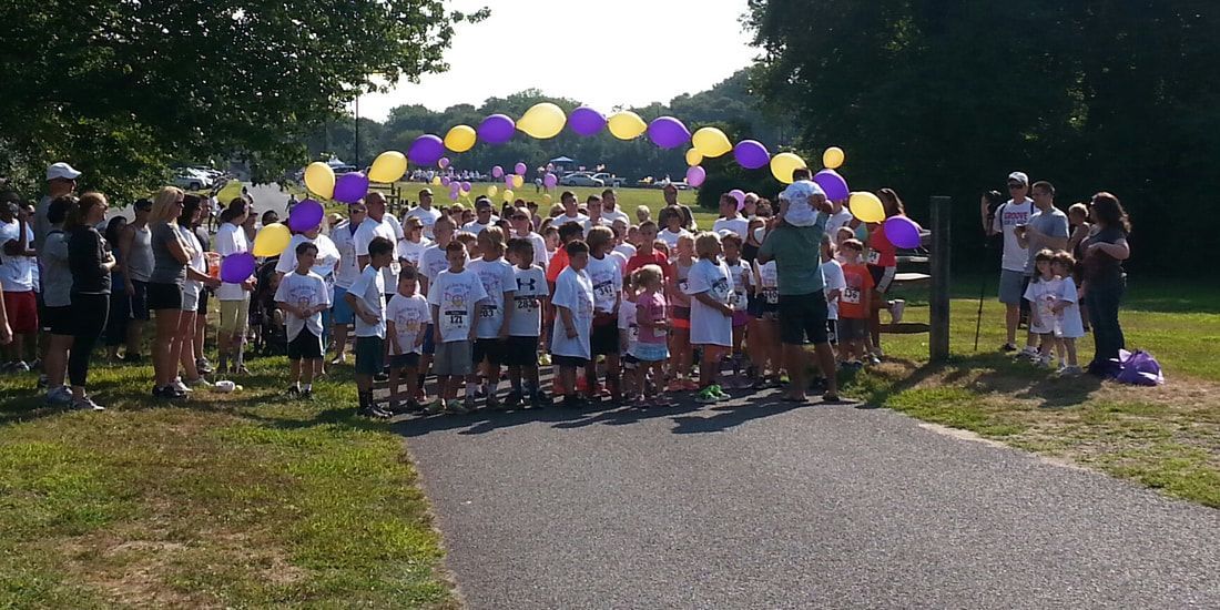 Group of people gathered for an outdoor event, some wearing white shirts, beneath balloons over a path.