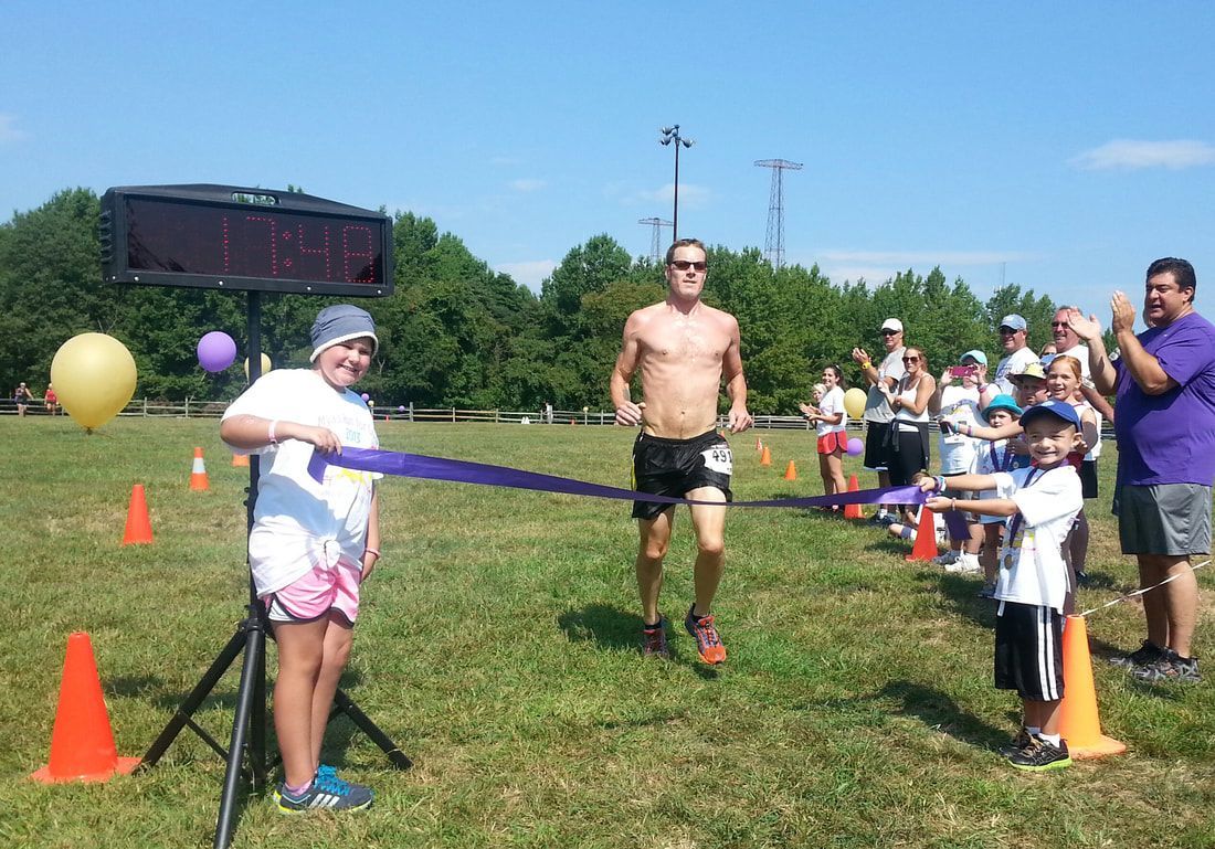 A shirtless runner breaks the finish line tape held by two children during an outdoor race.