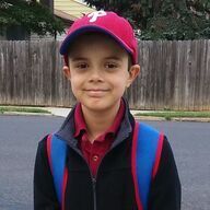 Boy in red Phillies cap, backpack, and jacket smiles in front of a fence and street.