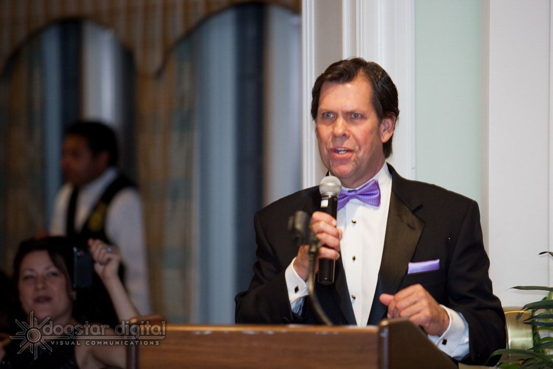 Man in tuxedo speaks at podium with purple bow tie; indoors.
