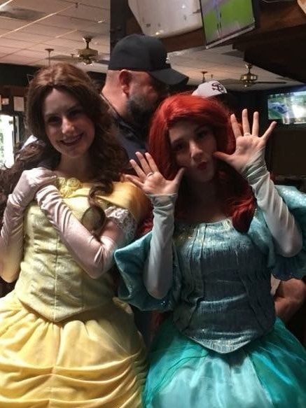 Two women in princess costumes, Belle and Ariel, smiling and posing in a bar.