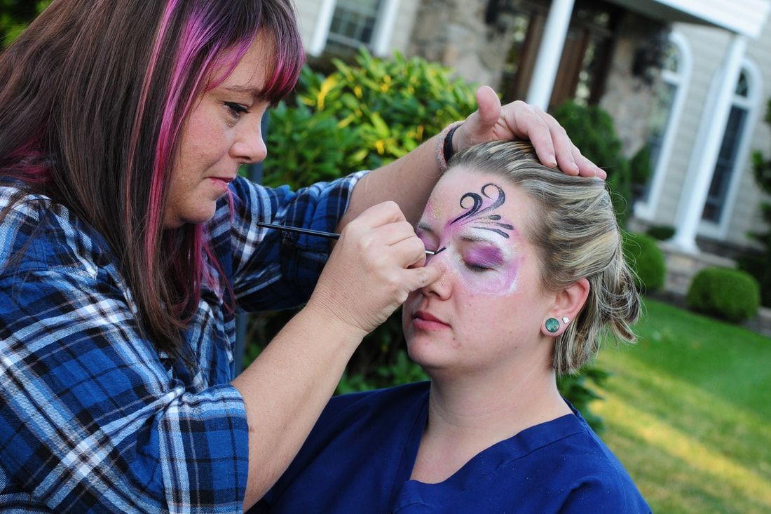 Woman painting another woman's face outdoors; purple and black design on forehead, pink hair streaks.
