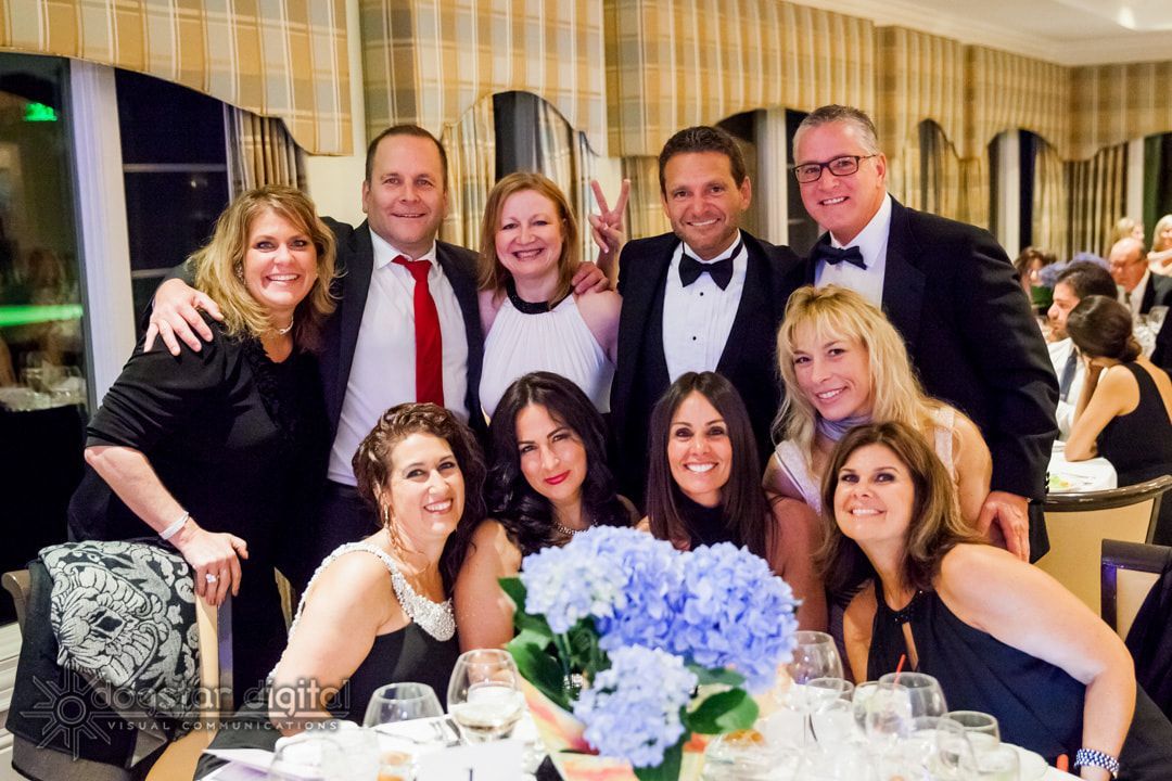 Group of people at a formal dinner, smiling and posing around a table with blue flowers.