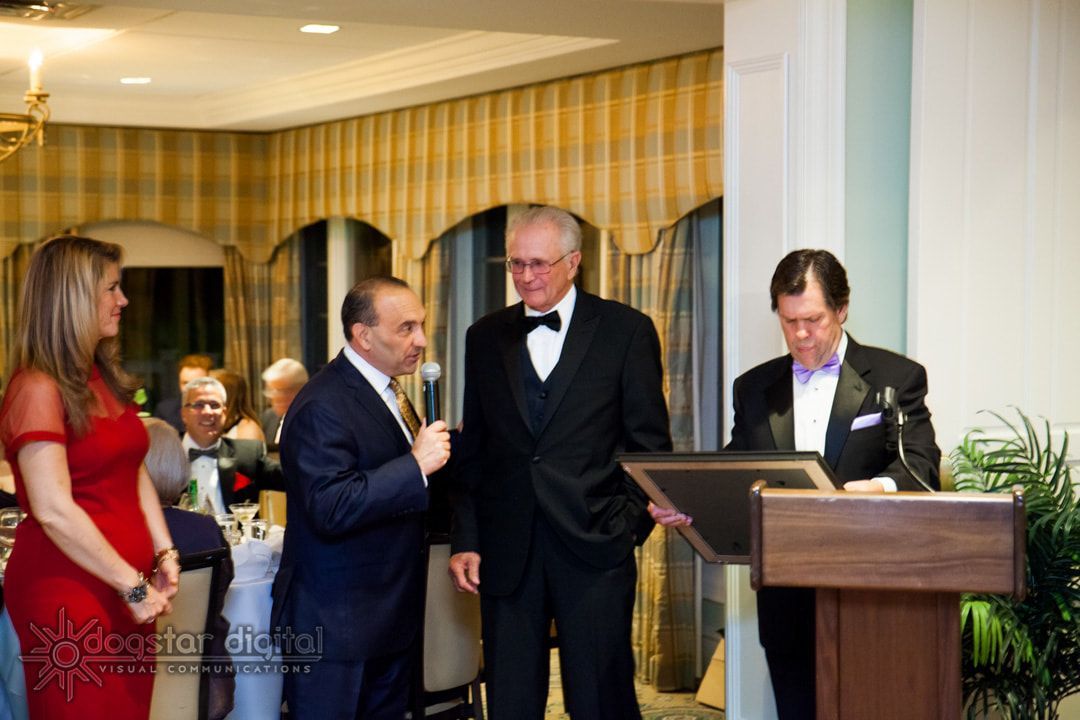 People at a formal event; man in tux holds a framed award; woman in red dress stands next to others at podium.