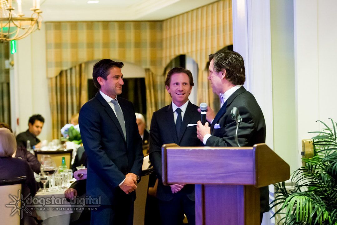 Three men in suits at a podium, one speaking with a microphone. Formal event, indoor setting.
