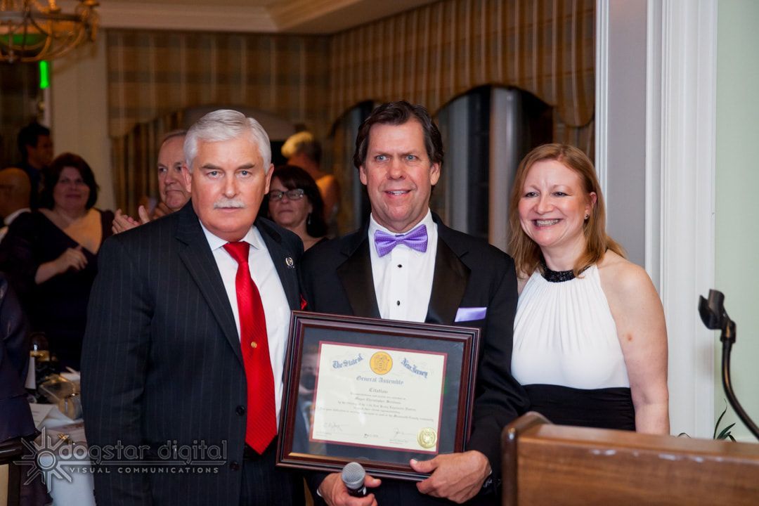 Three people at an event; man in tux holding framed certificate, flanked by people in formal attire.