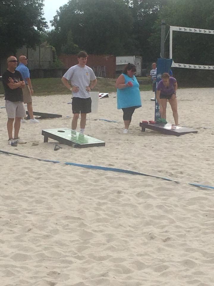 People playing cornhole on a sand court; one player throws, others watch.