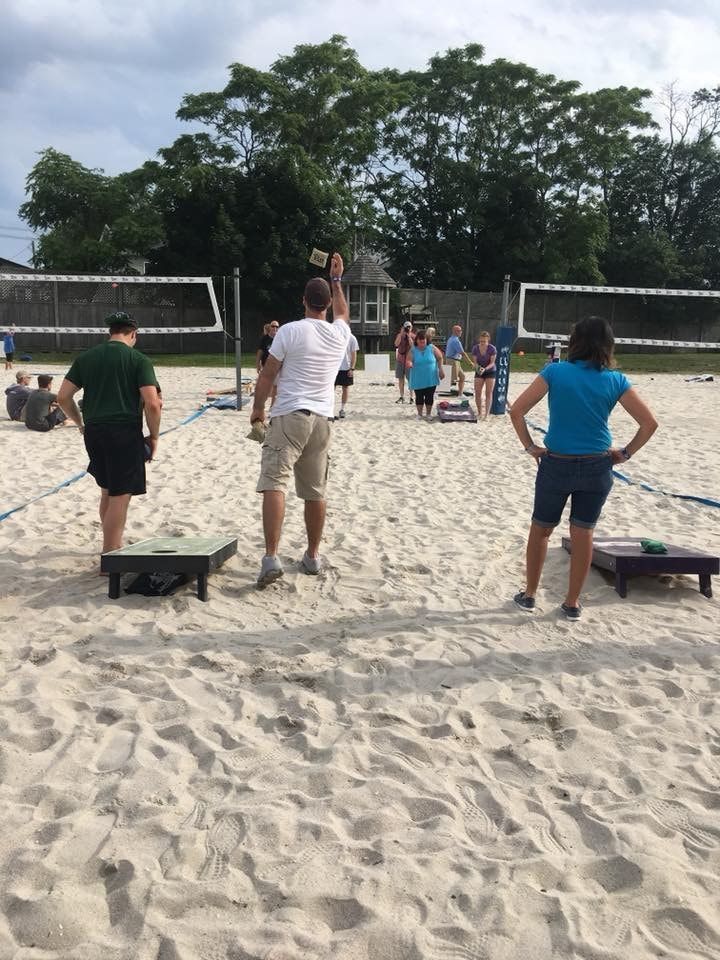People playing cornhole on a sandy court, trees in the background, cloudy sky.