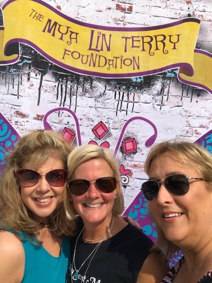 Three women smiling in front of a sign for The Mya Lin Terry Foundation. They are wearing sunglasses.
