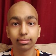 Bald-headed person with brown eyes looking at the camera against a red background. They are wearing a black and white shirt with orange.