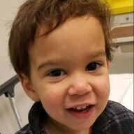 Young child with brown hair smiles, close-up. Plaid shirt. Indoor setting.