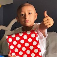 Boy with shaved head, holding a wrapped gift, giving a thumbs-up in a hospital bed.