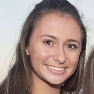 Woman with long brown hair smiles, outdoors with soft lighting.