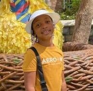 Person wearing a hat and yellow shirt smiles, in front of a yellow figure and nest.