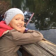Young person with a knitted hat smiles, leaning on wooden railing by water, sign in background.