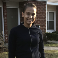 Woman in a black zip-up jacket smiles in front of a brick building.