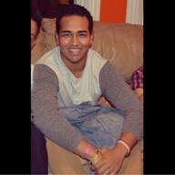 Man seated on a couch, wearing gray and white shirt, smiling.