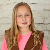 Young person with long blonde hair smiles, wearing a pink shirt and brown vest, against a white brick wall.