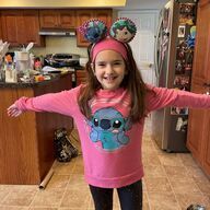 Girl with pink sweater and mouse ears smiles at camera, standing near a kitchen.