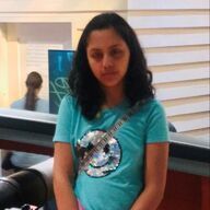 Woman in a turquoise shirt with a sequined smiley face smiles in an indoor setting.