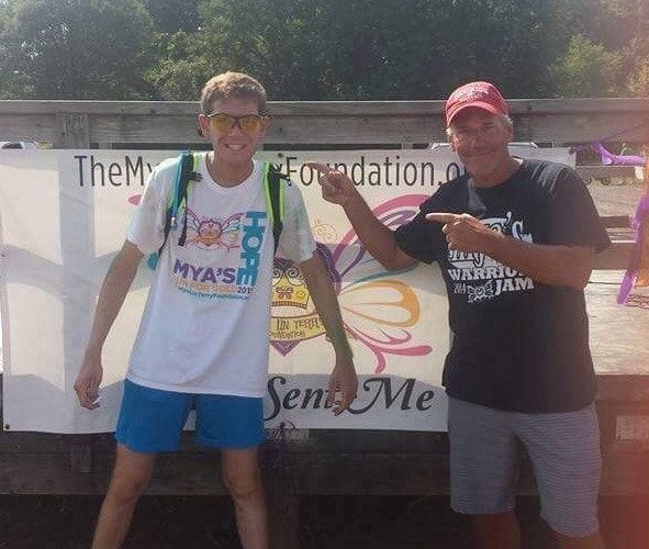 Two men standing in front of a banner. One points at the banner, the other at his shirt. Sunny, outdoor setting.