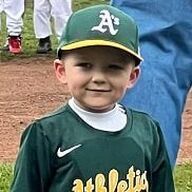 Boy in green baseball uniform and A's hat smiling, standing on a baseball field.