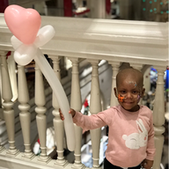 Young child with a balloon wand smiles in front of a white railing. The balloon is pink and white.