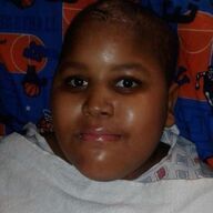 Person with smooth, dark skin, light eyes, and a slight smile, lying down with a medical gown and a blue patterned sheet.