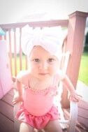 Baby in pink swimsuit and white bow headband sits in a white rocking chair on a porch.