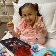 Girl in pink hoodie smiles in hospital bed, holding snack and pointing.