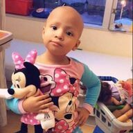 Child in Minnie Mouse pajamas, holding Minnie Mouse plush, with bald head, serious expression, in hospital room.