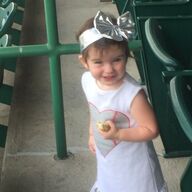 Child in a white shirt with heart design and silver bow headband, holding a snack near green stadium seats.