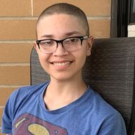 Person with shaved head wearing glasses, smiling, seated in a chair, wearing a blue Superman shirt.