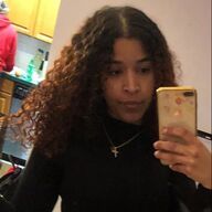 Woman with curly hair, in a black top, holding a phone, looking at the camera.