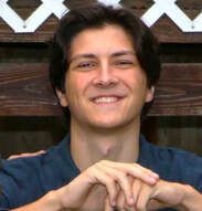 Young person smiling, hands clasped, wearing a dark blue shirt. Wooden background with a white trellis.