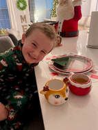 Smiling child in Christmas pajamas beside festive mugs and plates on a table.