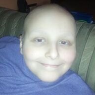 Person with a smooth, hairless head smiles; wearing a blue shirt, sitting on a couch.