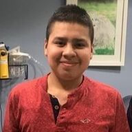 Boy with short dark hair smiles, wearing a red long-sleeved shirt, indoors near medical equipment.