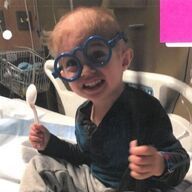 Smiling child wearing blue novelty glasses, holding a spoon, in a hospital bed.