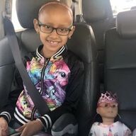 Girl in glasses, patterned jacket, smiles, seated in car next to doll wearing crown.