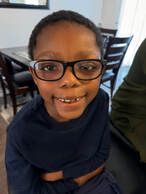 Boy wearing glasses smiles, arms crossed, indoor setting.