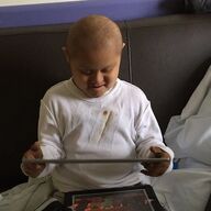 Child with short hair wearing a white shirt, using a tablet, seated on a bed.
