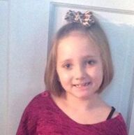 Girl with short hair, smiling, wearing a red top and a bow, standing in front of a white door.