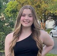 Woman with long wavy hair in a black one-shoulder top smiles outdoors in front of trees and a car.