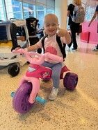 Child on a pink tricycle, smiling and giving a thumbs up. In a building with people and a stroller.