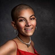 Woman with a shaved head smiles, wearing a red dress, pearl necklace, and hoop earrings, against a dark gray backdrop.